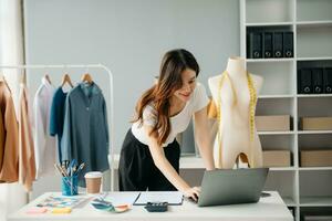 riuscito moda progettista. attraente giovane asiatico donna con Sorridi mentre in piedi nel officina. bellissimo proprietario attività commerciale donna Lavorando e detiene tavoletta, il computer portatile e smartphone nel studio. foto