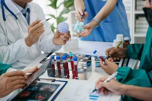 medico squadra avendo un' incontro con medici nel bianca laboratorio cappotti e chirurgico scrub seduto a un' tavolo discutere un' pazienti Lavorando in linea utilizzando computer nel il medico industria foto