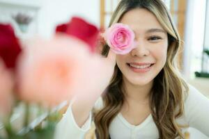 sorridente asiatico donna Tenere rosa fiore nel sua mano a casa foto