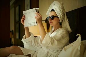 avvicinamento. attraente donna nel occhiali da sole, attività commerciale persona, avvolto nel bagno asciugamano dopo rilassante bagnarsi e godendo lettura giornale, seduta su un' letto nel Hotel Camera da letto durante sua attività commerciale viaggio foto