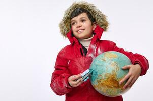 in giro il mondo viaggio e turismo concetto. adorabile elementare anziano scuola ragazzo nel caldo luminosa rosso giù giacca con cappuccio Tenere un aereo giocattolo modello e globo, imitando il volo in giro il terra foto