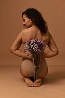 posteriore Visualizza di bellissimo africano donna nel biancheria intima, guardare per il lato, Tenere un' mazzo di fiori selvatici dietro a sua Indietro, seduta su beige sfondo. corpo positivo, accettazione di sé, femminilità concetto foto