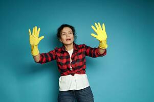 giovane ispanico donna guardare a sua mani nel giallo gomma da cancellare guanti per lavori di casa, fingendo per toccare e esaminare sporco su un invisibile superficie foto