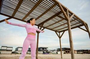 giovane sportivo donna in piedi con disteso braccia sotto un' di legno baldacchino su il spiaggia. all'aperto allenarsi concetto foto