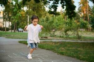 un attivo sportivo 4 anni vecchio bambino ragazza giochi campana , prende giri salto al di sopra di il piazze segnato su il terra. strada figli di Giochi nel classici. foto