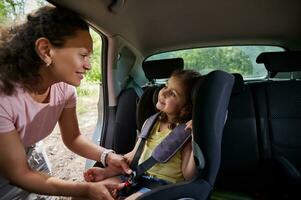 contento bambino ragazza sorrisi guardare a sua madre mentre lei è mettendo sua nel il auto posto a sedere cintura. sicuro movimento di bambini nel il auto foto