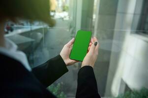 messa a fuoco su un' Telefono con un' verde croma chiave per inserire Annunci nel il mani di un irriconoscibile donna in piedi nel il finestra di un' grattacielo edificio prospiciente un urbano sfondo. copia spazio. foto