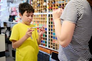 grave adolescenziale ragazzo sceglie acquerello marcatori nel scuola Stazionario negozio, chiede un' i saldi manager per Aiuto foto