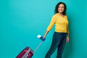 contento Riccio dai capelli scuri donna, viaggiatore nel luminosa giallo maglione e blu jeans Tenere passaporto con imbarco passaggio e aria biglietto, sorrisi, in posa con bagaglio su blu sfondo con copia spazio foto