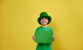 sorridente ragazzo indossare verde irlandesi fata cappello pose spirito un' foglio di verde carta nel il suo mani. festeggiare santo Patrick giorno concetto. giallo sfondo. copia spazio. foto