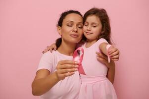 adorabile razza mista donna madre abbracci un' poco ragazza nel rosa abiti e viene visualizzato un' rosa raso nastro per telecamera, simbolo di Seno cancro giorno nel ottobre mese. supporto per cancro pazienti e sopravvissuti foto