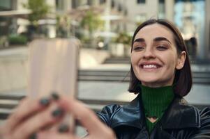 avvicinamento stile di vita ritratto di bellissimo divertente giovane brunetta con elegante corto capelli, Tenere smartphone nel disteso mani e sorridente trentadue denti Sorridi assunzione autoscatto mentre in piedi all'aperto nel città foto