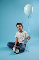 simpatico ragazzo sorrisi seduta vicino un allarme orologio su un' blu sfondo e Tenere un' blu Palloncino. copia spazio. festeggiare figli di giorno. foto