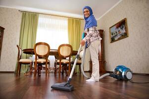 delizioso contento Arabo musulmano giovane donna o casalinga con vuoto più pulito, passare l'aspirapolvere il pavimento a casa. domestico, lavori di casa e pulizia concetto. foto