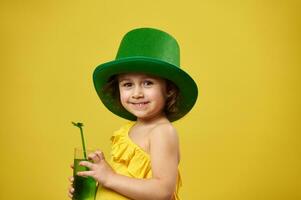 carino poco ragazza indossa un' fata irlandesi verde cappello sorrisi per il telecamera con un' bicchiere di verde bevanda nel sua mani. santo Patrick giorno. foto