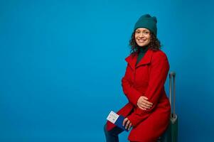 delizioso donna nel rosso cappotto, verde cappello e Maglione, con passaporto, aria biglietto, imbarco passaggio, si siede su valigia e sorrisi trentadue denti Sorridi guardare a telecamera. viaggio. turismo concetto. blu sfondo foto