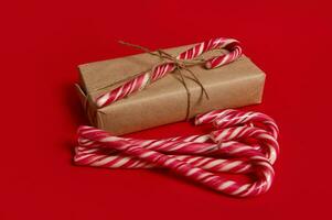 bellissimo minimalista composizione di Natale Oggetti, regalo scatola avvolto nel kraft involucro regalo carta con legato arco e dolce caramella canne isolato al di sopra di rosso sfondo con copia spazio per annuncio pubblicitario foto