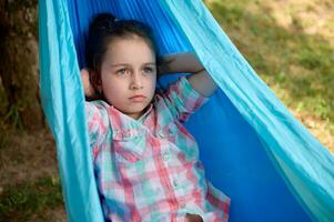 avvicinamento bello poco bambino ragazza rilassante su blu amaca nel il Giardino dietro la casa, godendo un' fine settimana all'aperto. bambini. svaghi foto