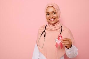 bellissima arabo musulmano femmina medico con bellissimo trentadue denti Sorridi mostrando un' rosa raso nastro, isolato su colorato sfondo con copia spazio. mondo giorno di combattimento Seno cancro, 1 st ottobre concetto foto