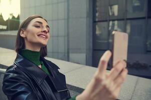 avvicinamento stile di vita ritratto di allegro divertente giovane brunetta con elegante corto capelli, Tenere mobile Telefono nel disteso mani e sorridente trentadue denti Sorridi assunzione autoscatto mentre in piedi all'aperto nel città foto