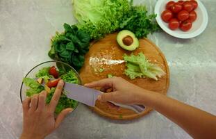 avvicinamento di femmina capocuoco mani con un' cucina coltello affettare pomodori e lancio loro in un' bicchiere trasparente arco mentre preparazione salutare crudo vegano insalata. alto angolo Visualizza foto