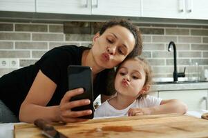 madre e figlia guardare a mobile Telefono telecamera e assunzione autoscatto nel il cucina durante cibo preparazione foto