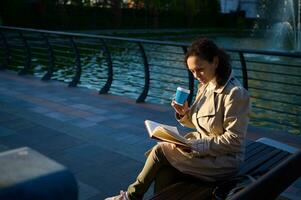 giovane donna nel beige trincea cappotto seduta su un' parco panchina su il lago sfondo , potabile caffè o caldo bevanda nel riciclabile porta via carta tazza e lettura prenotare, godendo riposo a partire dal digitale gadget foto