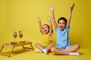 Due adorabile bambini seduta nel loto posizione vicino un' di legno servendo vassoio con cocktail e piatto con affettato frutta e raccolta braccia su, ridendo e esprimendo felicità foto