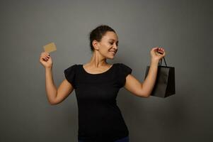 bella sorridente africano donna vestito nel nero detiene un' shopping Borsa e oro credito carta, Spettacoli loro in posa per telecamera, isolato al di sopra di grigio parete sfondo con copia spazio per nero Venerdì annuncio pubblicitario foto