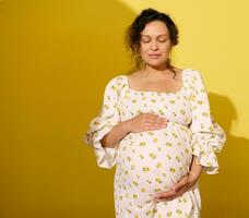 autentico incinta donna Tenere sua gonfiarsi, vestito nel estate vestire, in posa con occhi chiuso su giallo studio sfondo foto