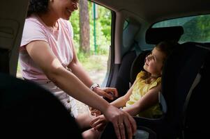 sorridente donna con sua carino figlia guardare a ogni altro mentre madre fissaggio sua figlia con cintura di sicurezza su il bambino auto posto a sedere foto