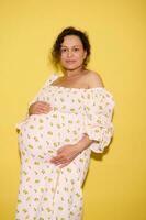 incinta donna nel in ritardo gravidanza delicatamente colpi sua grande gonfiarsi, guardare con fiducia a telecamera, isolato giallo sfondo foto
