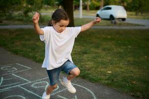 avvicinamento di poco caucasico ragazza giocando campana su asfalto. bambino giocando campana gioco su terreno di gioco all'aperto su un' soleggiato giorno. strada figli di Giochi nel classici. foto