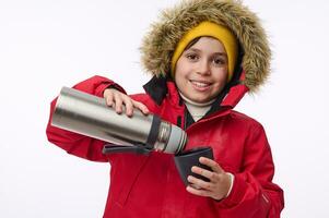 orizzontale ritratto di un' carino età scolastica ragazzo inverno viaggiatore avventuriero scrosciante caldo bevanda a partire dal thermos in boccale sorridente trentadue denti Sorridi guardare a telecamera isolato su bianca sfondo con copia spazio foto