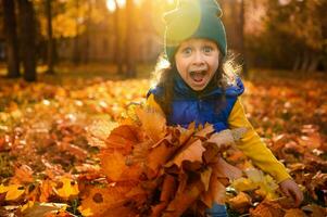 emotivo stile di vita ritratto di adorabile allegro bambino ragazza nel colorato Abiti giocando con asciutto caduto autunno acero le foglie nel d'oro parco a tramonto con bellissimo raggi di sole caduta attraverso alberi foto