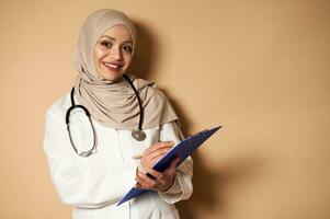 fiducioso ritratto di arabo musulmano medico nel hijab in piedi con appunti nel mani su beige sfondo e sorridente trentadue denti Sorridi guardare a telecamera foto