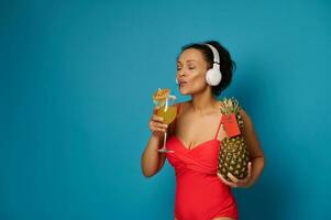 misto gara donna nel rosso costume da bagno detiene ananas con vuoto rosso carta e bevande esotico cocktail mentre godendo musica con cuffie foto