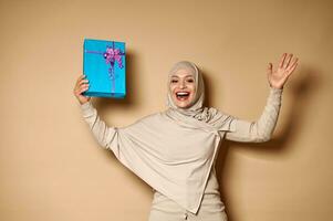 contento musulmano donna nel hijab raccolta sua mani con un' regalo, esprimendo felicità per telecamera. beige sfondo, copia spazio. foto