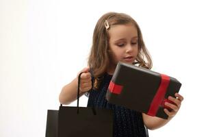 ritratto di un' bellissimo 4 anni vecchio poco ragazza con chiuso occhi Tenere un' regalo e shopping pacchetto, in posa contro un' bianca sfondo con copia spazio. nero Venerdì concetto foto