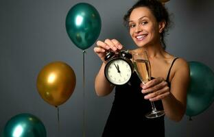 bellissimo donna nel sera nero vestito detiene un' flauto di scintillante vino e Spettacoli un allarme orologio isolato al di sopra di grigio sfondo con gonfiato aria palloncini. Natale e nuovo anno concetto, anniversario. foto