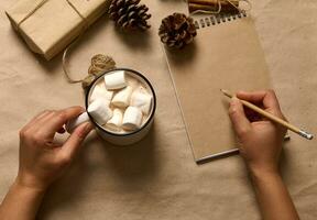 piatto posizione. umano mani hold un' tazza con caldo cacao bevanda e marshmallows e scrittura con di legno matita su vuoto carta foglio di bloc notes su il cartone superficie con mestiere Natale regalo e pino coni foto