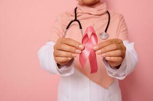 avvicinamento di mano di medico nel rosa hijab, Tenere rosa nastro. mondo cancro giorno. Seno e addominale cancro consapevolezza, ottobre rosa giorno, colorato sfondo, copia spazio. Seno cancro supporto concetto. foto