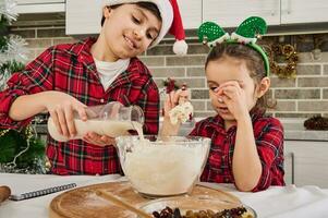 bello preadolescente caucasico ragazzo vestito nel rosso e verde scacchi camicia e Santa cappello versa alcuni latte in un' ciotola, cucinando con il suo poco sorella Natale torta a casa cucina. foto