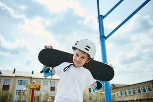 adorabile ragazzo nel un' protettivo casco e attrezzatura in posa con un' skateboard su il suo le spalle dietro a il suo testa foto