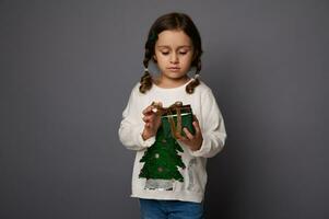 adorabile ragazza nel bianca maglione con brillante Natale albero detiene un' regalo scatola avvolto nel brillante verde involucro carta isolato su grigio sfondo con copia spazio. nuovo anno Natale concetto per pubblicità foto