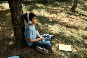 contento ispanico adolescenziale ragazzo nel cuffia, rilassante nel il parco dopo scuola, seduta sotto albero vicino sparpagliato libri di testo foto