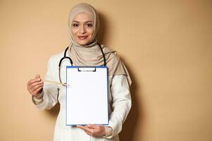 affascinante femmina medico nel hijab puntamento con un' matita a un' vuoto foglio di carta su un' clipboard. assistenza sanitaria e assicurazione concetto. copia spazio per promozione foto