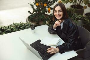 alto Visualizza di libero professionista donna, giovane attività commerciale persona, copywriter, carino sorridente guardare a telecamera mentre seduta a tavolo con un' bloc notes e computer portatile, Lavorando in linea remoto nel un' terrazza di caffetteria foto