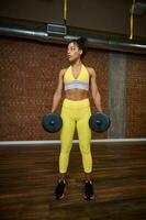 bellissimo africano donna con salutare sottile corpo e piatto pancia nel giallo tuta da ginnastica esercizio con manubri . corpo cura, peso perdita concetto foto