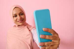 morbido messa a fuoco su il smartphone nel blu copertina nel disteso braccia di arabo musulmano donna indossare tradizionale religioso islamico vestito, rosa hijab e sorrisi con trentadue denti Sorridi fabbricazione auto ritratto, autoscatto foto